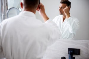Combing hair. Close up photo of a calm man wearing a bathrobe and looking attentively at his reflection in the mirror while combing his hair